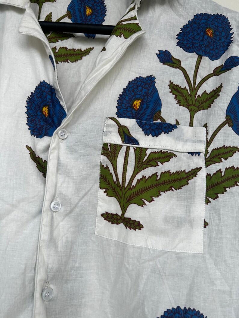 May include: White button-down shirt with a floral pattern. The shirt features large blue flowers with yellow centers and green leaves. A pocket on the chest displays a matching floral design. The shirt is made of a lightweight fabric.