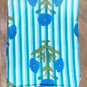 May include: Folded fabric with a striped pattern in shades of turquoise and white. The fabric features a repeating floral design in blue and green, with stylized flowers and foliage. The fabric is folded neatly, showing the pattern clearly.
