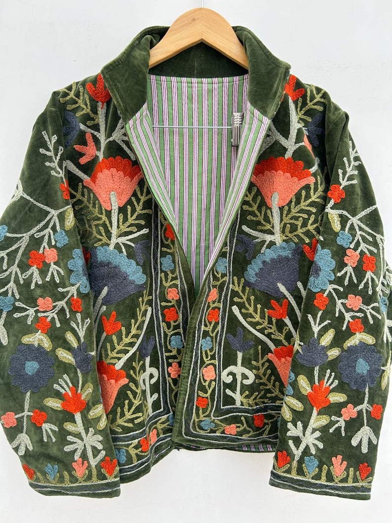 May include: A green velvet jacket with intricate floral embroidery in shades of red, blue, and pink. The jacket features a striped lining in green, pink, and white. The jacket is hanging on a wooden hanger.