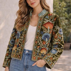Könnte beinhalten: Olivgrüne Samtjacke mit farbenfroher Blumenstickerei. Die Jacke hat einen Stehkragen mit grün-weißen Streifen und einen Knopfverschluss. Die Ärmel sind lang und die Jacke wird mit einer blauen Jeans getragen.