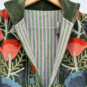 May include: A dark green velvet jacket with floral embroidery in shades of orange, blue, and green. The jacket features a striped lining in green and white with pink accents and a green velvet collar.