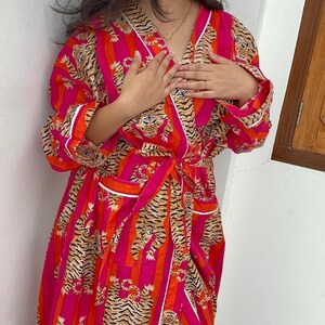 May include: A vibrant, knee-length robe in a bold pink hue, featuring vertical orange stripes and a repeating tiger print. The robe has a V-neck, long sleeves, and a matching tie belt. White trim accents the edges and pockets, creating a striking contrast.
