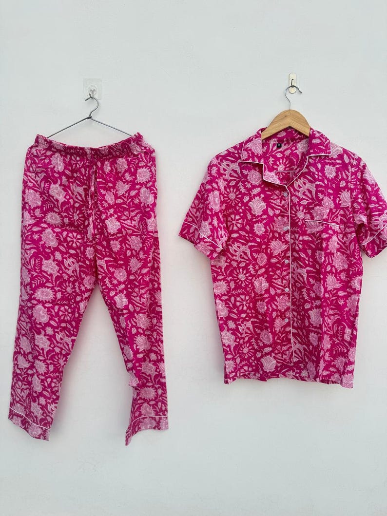 May include: A two-piece pajama set in a vibrant fuchsia color, featuring a floral pattern in a lighter shade of pink. The set includes a short-sleeved button-down shirt and matching drawstring pants. The shirt has white piping along the collar and cuffs.