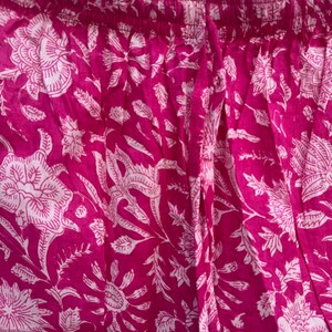 May include: Close-up of a pair of pink pants with a white floral pattern. The fabric is a vibrant fuchsia color, and the floral design includes large, stylized flowers and leaves. The pants have an elastic waistband.