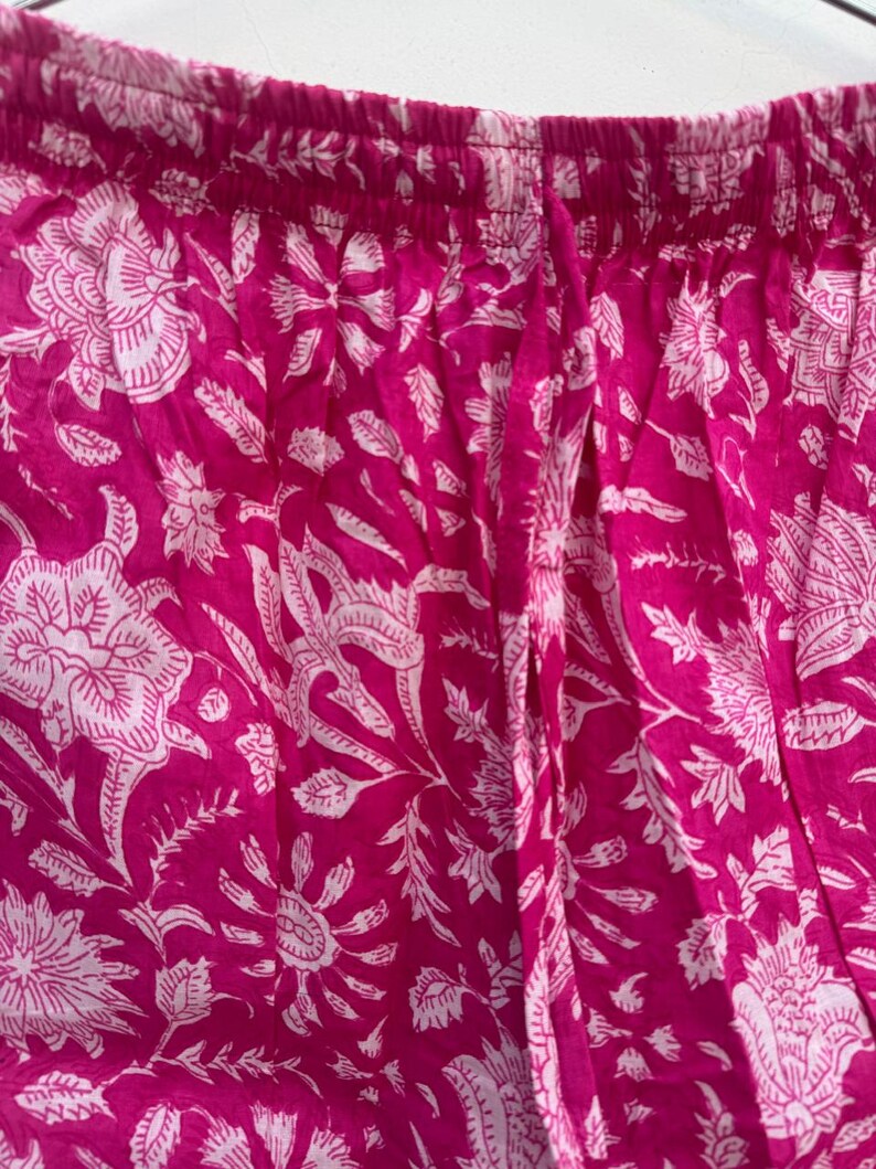 May include: Close-up of a pair of pink pants with a white floral pattern. The fabric is a vibrant fuchsia color, and the floral design includes large, stylized flowers and leaves. The pants have an elastic waistband.
