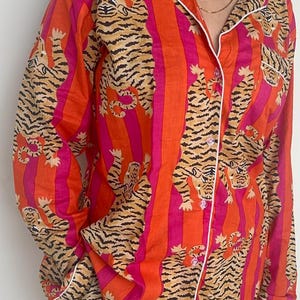 May include: A two-piece pajama set with a tiger print. The set includes a button-down shirt and matching trousers. The shirt and trousers have a striped pattern in orange and pink, with a repeating tiger illustration.