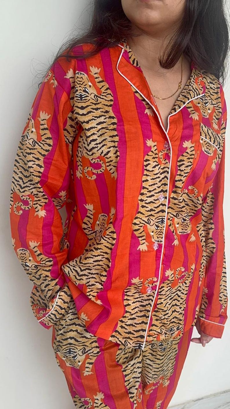 May include: A two-piece pajama set with a tiger print. The set includes a button-down shirt and matching trousers. The shirt and trousers have a striped pattern in orange and pink, with a repeating tiger illustration.