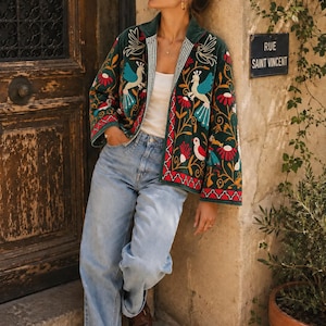 May include: A dark green velvet jacket featuring vibrant multi-colored embroidery of birds and floral patterns, worn over a white tank top and light blue denim jeans. The jacket has long sleeves, a straight hem, and a striped inner lining detail.