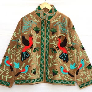May include: Brown jacket with embroidered birds in red, black, and turquoise. The jacket features a high collar and a zipper closure. Green embroidered vines and accents frame the birds. The jacket is hanging on a wooden hanger.