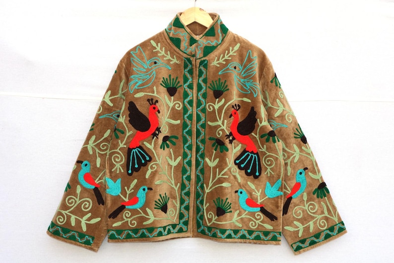 May include: Brown jacket with embroidered birds in red, black, and turquoise. The jacket features a high collar and a zipper closure. Green embroidered vines and accents frame the birds. The jacket is hanging on a wooden hanger.
