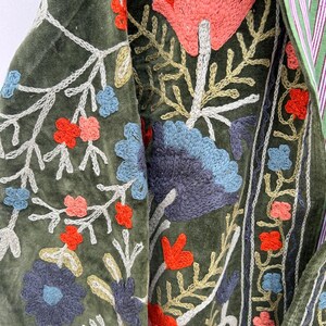 May include: Close-up of a green velvet garment with intricate floral embroidery. The embroidery features red, blue, and peach-colored flowers with white and gold accents. The garment has a striped green and white lining.