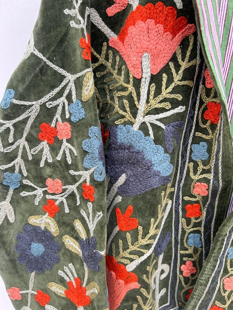May include: Close-up of a green velvet garment with intricate floral embroidery. The embroidery features red, blue, and peach-colored flowers with white and gold accents. The garment has a striped green and white lining.