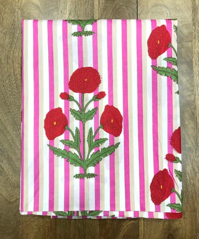 May include: Folded fabric with a pink and white striped pattern. Large red poppy flower designs with green leaves are printed on the fabric. The fabric is folded neatly, showcasing the floral and striped design.