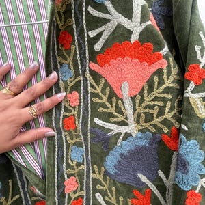 May include: Olive green jacket with embroidered floral design in shades of red, blue, and peach. The jacket features a striped lining in green and white. The embroidery is detailed and covers the front of the jacket.