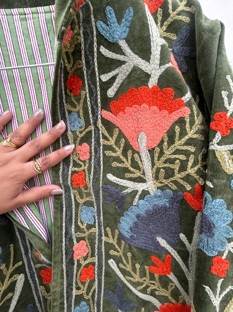 May include: Olive green jacket with embroidered floral design in shades of red, blue, and peach. The jacket features a striped lining in green and white. The embroidery is detailed and covers the front of the jacket.