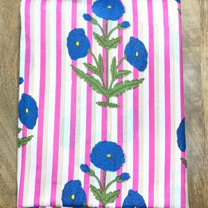 May include: Folded fabric with a pink and white striped background. The fabric features a repeating pattern of blue flowers with green leaves and stems. The flowers have yellow centers.