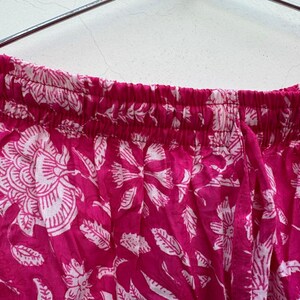 May include: Close-up of a section of pink fabric with a white floral pattern. The fabric is gathered at the top, suggesting it's part of a garment like pants or a skirt. The floral design includes large flowers and leaf shapes.