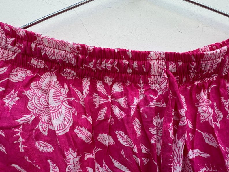 May include: Close-up of a section of pink fabric with a white floral pattern. The fabric is gathered at the top, suggesting it's part of a garment like pants or a skirt. The floral design includes large flowers and leaf shapes.