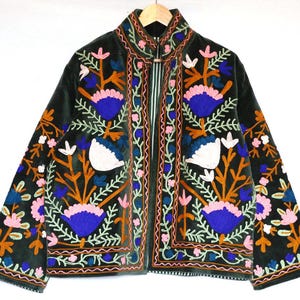 May include: Dark green velvet jacket with colorful floral embroidery. The jacket features a high collar and is adorned with orange, blue, pink, and white flower designs. The jacket is hanging on a wooden hanger.
