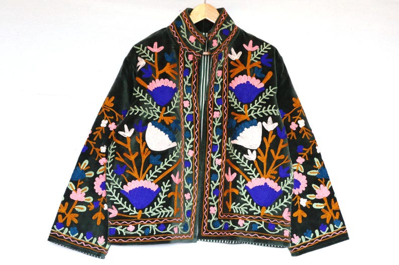 May include: Dark green velvet jacket with colorful floral embroidery. The jacket features a high collar and is adorned with orange, blue, pink, and white flower designs. The jacket is hanging on a wooden hanger.