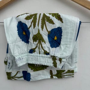 May include: Light blue pajama pants with a floral pattern. The design features large blue flowers with yellow centers and green leaves. The pants are folded and hanging on a wooden hanger.