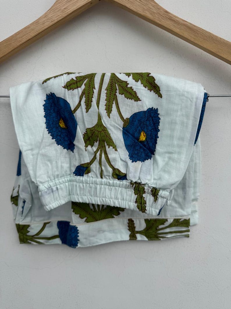 May include: Light blue pajama pants with a floral pattern. The design features large blue flowers with yellow centers and green leaves. The pants are folded and hanging on a wooden hanger.