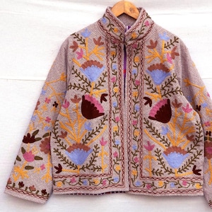 May include: A tan jacket with intricate floral embroidery in shades of yellow, pink, blue, and brown. The jacket features a high collar, long sleeves, and a full zipper closure. The embroidery is detailed and covers the front, sleeves, and collar.