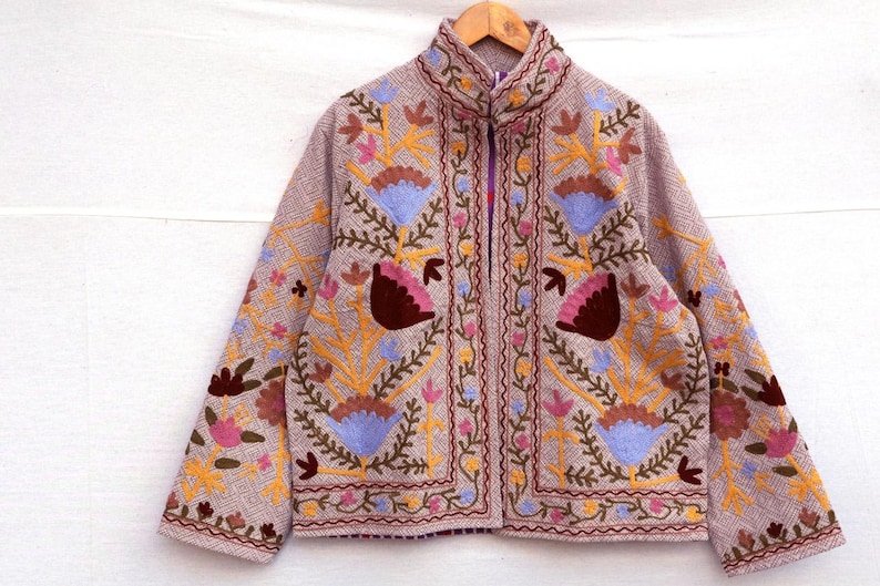 May include: A tan jacket with intricate floral embroidery in shades of yellow, pink, blue, and brown. The jacket features a high collar, long sleeves, and a full zipper closure. The embroidery is detailed and covers the front, sleeves, and collar.