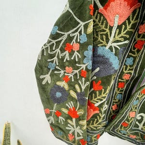 May include: Olive green velvet jacket with intricate floral embroidery in shades of red, blue, and peach. The embroidery features detailed patterns and a contrasting white outline, creating a textured and vibrant design. The jacket's design is visible on the sleeves and front.