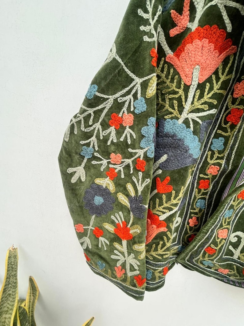 May include: Olive green velvet jacket with intricate floral embroidery in shades of red, blue, and peach. The embroidery features detailed patterns and a contrasting white outline, creating a textured and vibrant design. The jacket's design is visible on the sleeves and front.