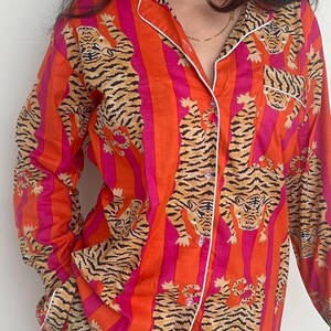 May include: Orange and pink striped pyjama set with a tiger print. The long-sleeved top has white trim and buttons. The matching trousers are also visible. The pyjamas are made of a soft material.