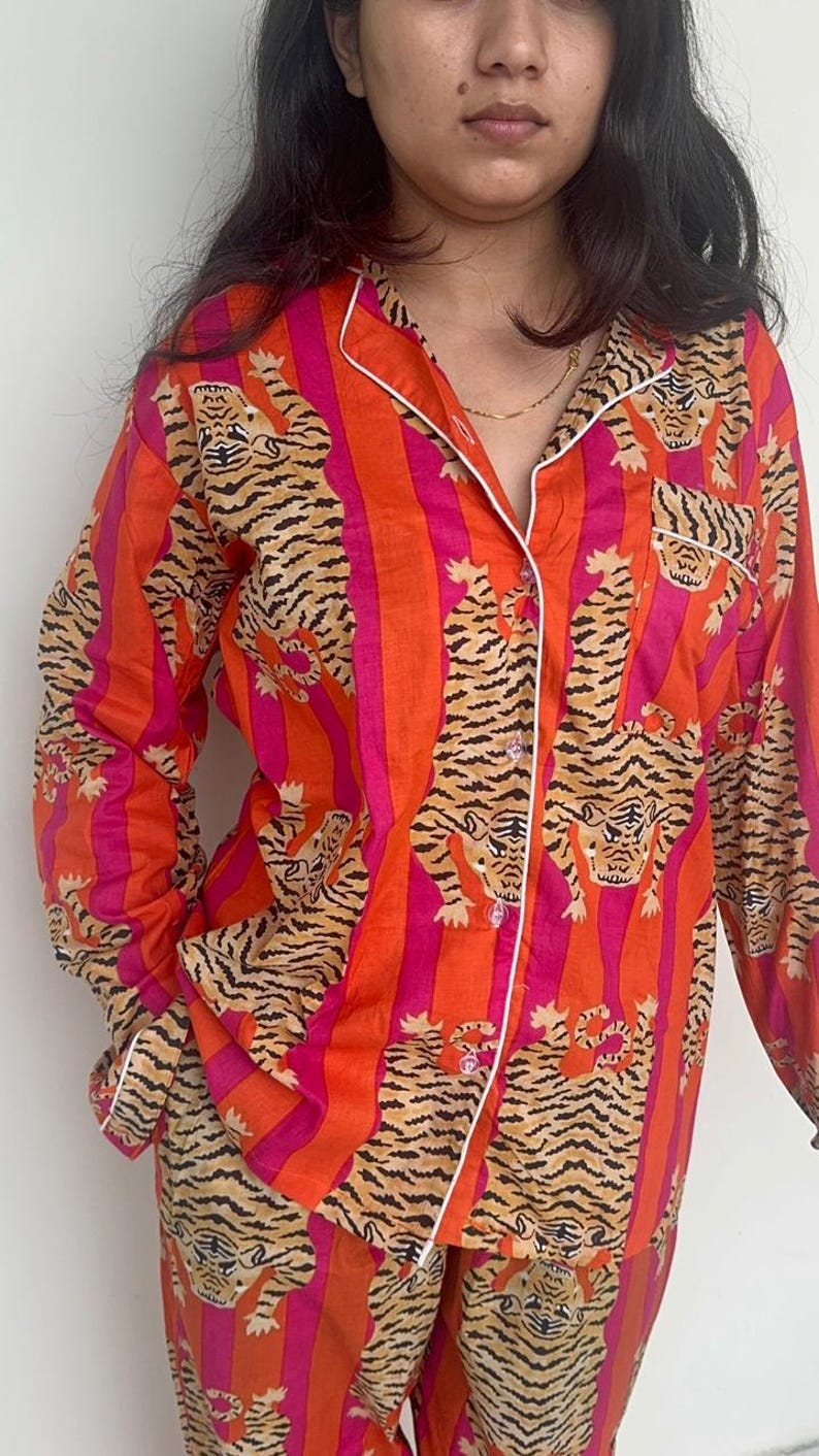 May include: Orange and pink striped pyjama set with a tiger print. The long-sleeved top has white trim and buttons. The matching trousers are also visible. The pyjamas are made of a soft material.