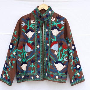 May include: Brown jacket with floral embroidery in white, red, and blue. The jacket has a stand-up collar and a full-length zipper. The floral design is repeated on the sleeves and front panels. The jacket is hanging on a wooden hanger.