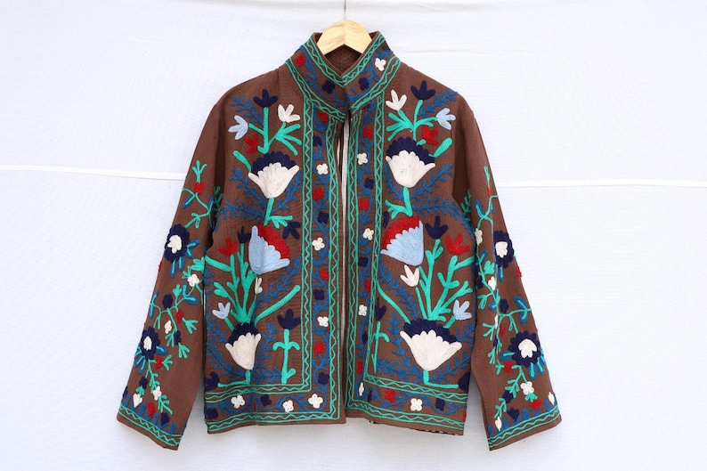 May include: Brown jacket with floral embroidery in white, red, and blue. The jacket has a stand-up collar and a full-length zipper. The floral design is repeated on the sleeves and front panels. The jacket is hanging on a wooden hanger.