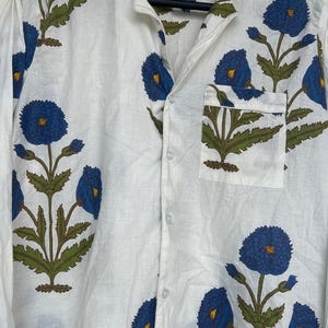May include: White button-down shirt with a blue floral pattern. The shirt features a classic collar, a chest pocket, and long sleeves. The floral design includes blue flowers with yellow centers and green leaves.