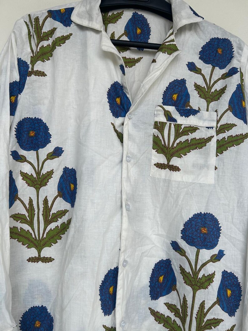 May include: White button-down shirt with a blue floral pattern. The shirt features a classic collar, a chest pocket, and long sleeves. The floral design includes blue flowers with yellow centers and green leaves.