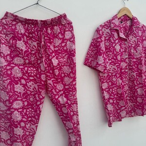 May include: A two-piece pajama set in a vibrant fuchsia hue, featuring a floral pattern in white. The set includes a button-down, short-sleeved shirt and matching pants. The pajamas are hanging on a hanger against a white background.