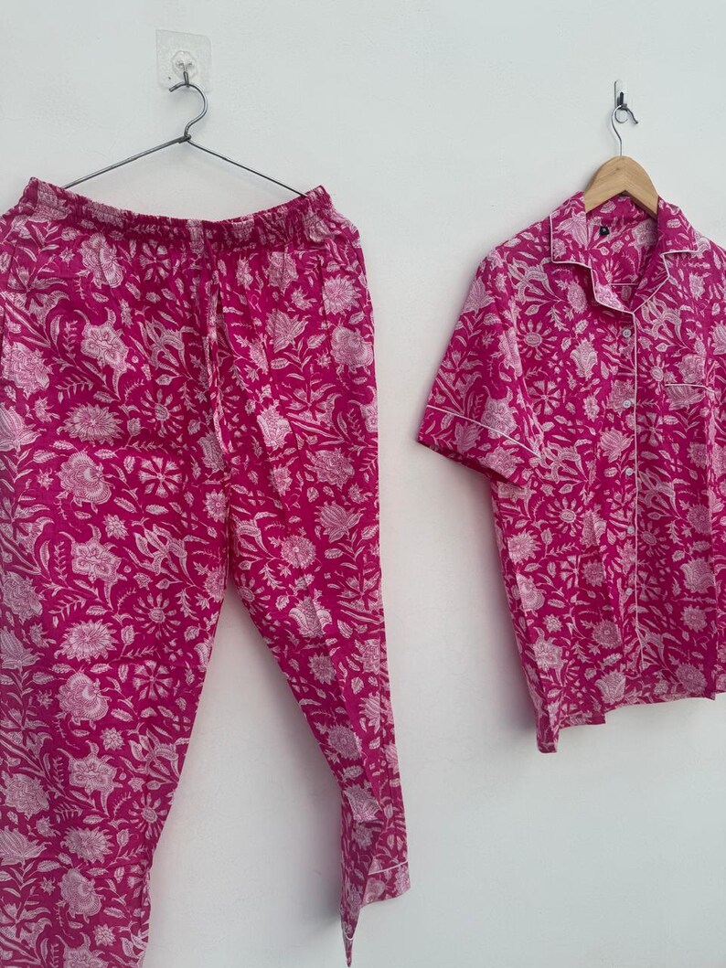 May include: A two-piece pajama set in a vibrant fuchsia hue, featuring a floral pattern in white. The set includes a button-down, short-sleeved shirt and matching pants. The pajamas are hanging on a hanger against a white background.