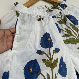 May include: White fabric garment with a blue floral print. The design features large blue flowers with yellow centers and green leaves. The garment is hanging on a wooden hanger.