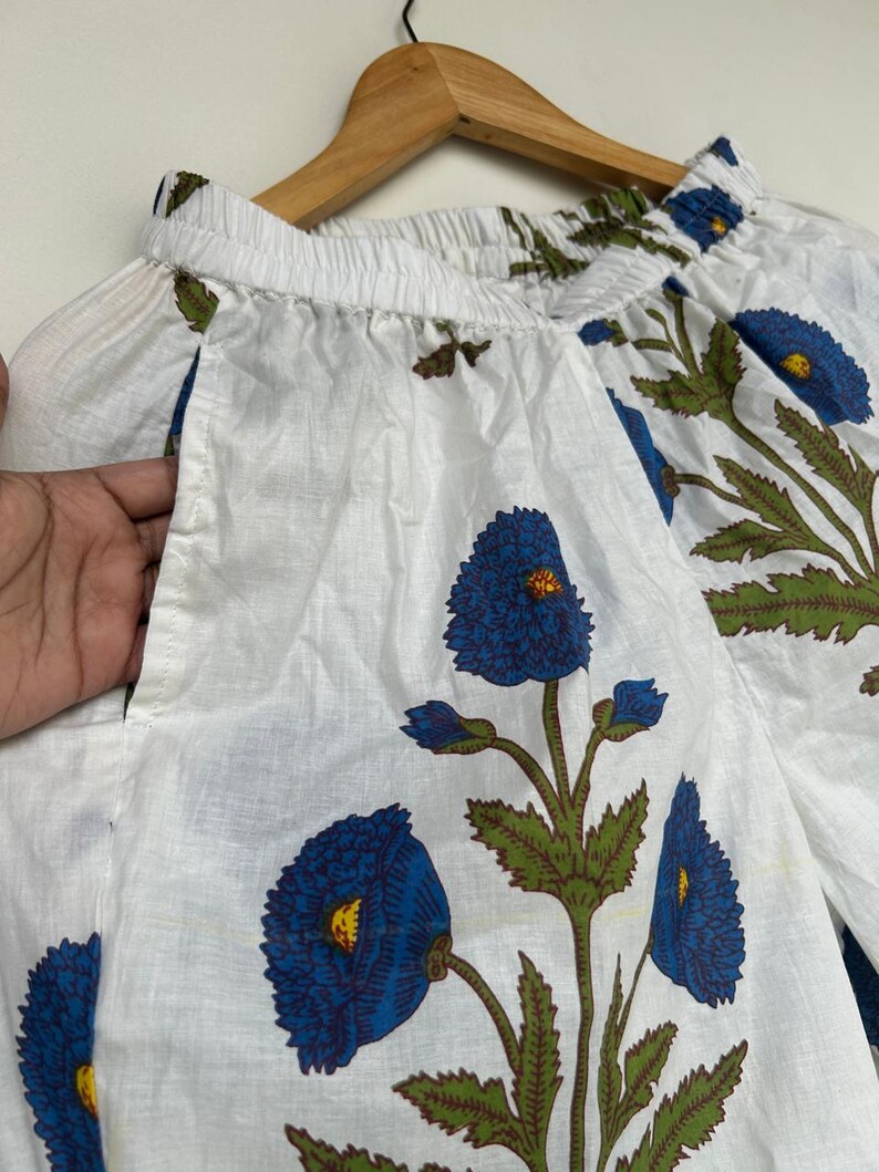 May include: White fabric garment with a blue floral print. The design features large blue flowers with yellow centers and green leaves. The garment is hanging on a wooden hanger.
