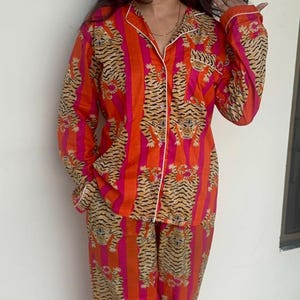 May include: An eye-catching orange pajama set featuring a tiger stripe design. The set comprises a button-down shirt and matching trousers, both trimmed with white. The pajamas are made from a soft, lightweight material.