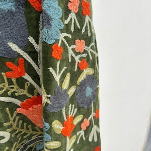 May include: Close-up of a green fabric with intricate floral embroidery. The embroidery features a variety of colors, including orange, blue, and gray, creating a detailed and textured design. The floral pattern is set against a dark green background.