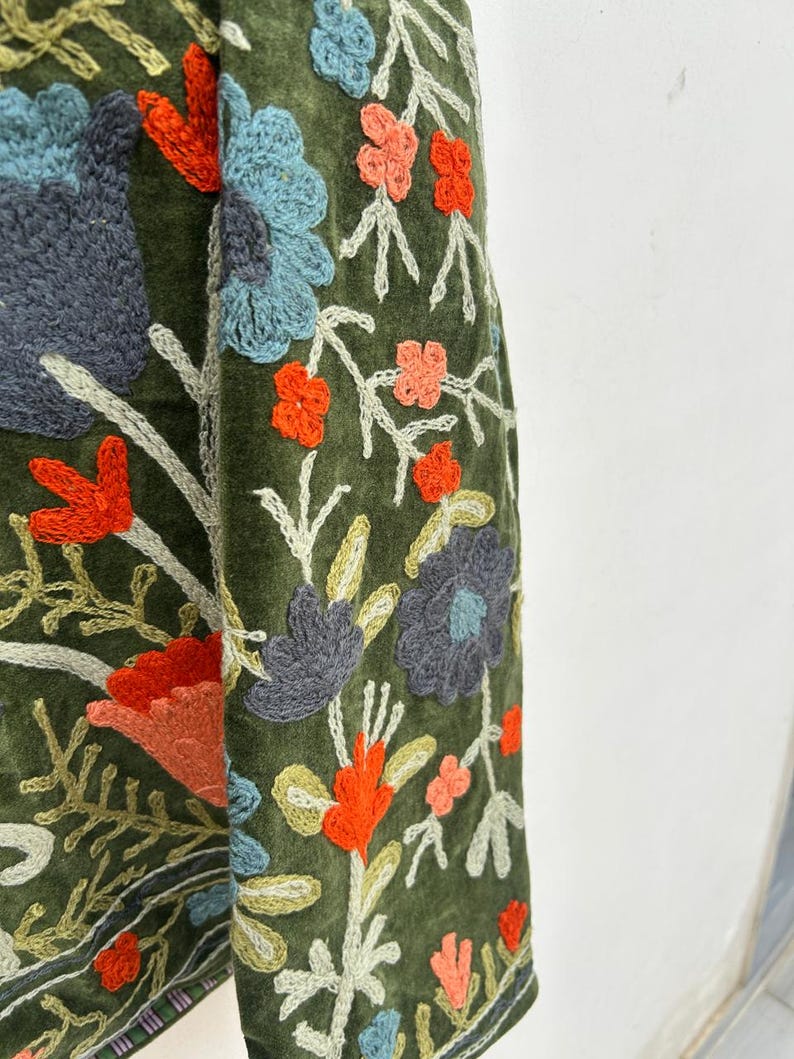 May include: Close-up of a green fabric with intricate floral embroidery. The embroidery features a variety of colors, including orange, blue, and gray, creating a detailed and textured design. The floral pattern is set against a dark green background.