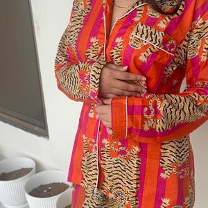 May include: A two-piece pyjama set in orange and pink stripes, featuring a tiger print. The long-sleeved top has white trim and a breast pocket. The matching trousers complete the sleepwear set.