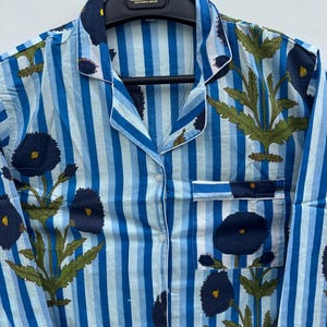 May include: Blue and white striped pajama top with a floral pattern. The top features a button-down front, a chest pocket, and white trim. The floral design includes dark blue flowers and green leaves. The garment is hanging on a black hanger.