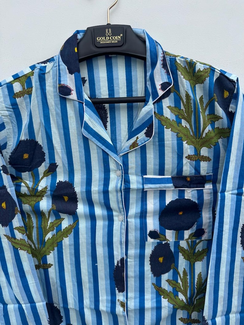May include: Blue and white striped pajama top with a floral pattern. The top features a button-down front, a chest pocket, and white trim. The floral design includes dark blue flowers and green leaves. The garment is hanging on a black hanger.