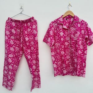 May include: A two-piece pajama set in a vibrant fuchsia hue, adorned with a delicate floral pattern in a lighter shade of pink. The set includes a button-down short-sleeved shirt with white trim and matching drawstring pants. The pajamas are displayed on hangers against a white background.