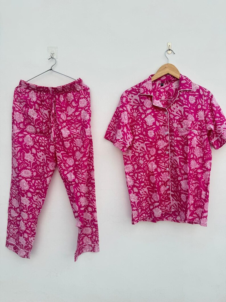 May include: A two-piece pajama set in a vibrant fuchsia hue, adorned with a delicate floral pattern in a lighter shade of pink. The set includes a button-down short-sleeved shirt with white trim and matching drawstring pants. The pajamas are displayed on hangers against a white background.