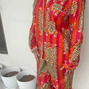 May include: A vibrant orange and pink striped pyjama set with a tiger print. The long-sleeved top and trousers display a repeating tiger and stripe pattern. White trim details the collar, cuffs, and pockets.
