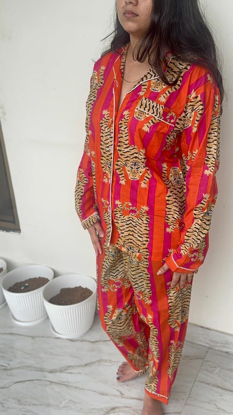May include: A vibrant orange and pink striped pyjama set with a tiger print. The long-sleeved top and trousers display a repeating tiger and stripe pattern. White trim details the collar, cuffs, and pockets.